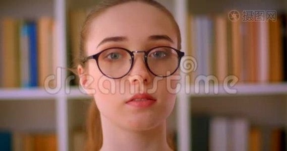 年轻漂亮的红头发女学生戴着眼镜，在室内看着图书馆里的摄像机视频