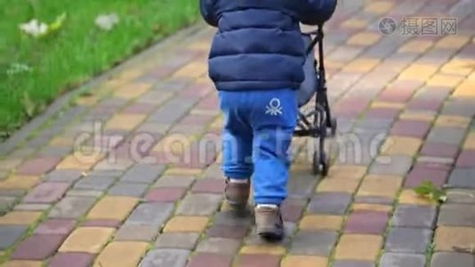 可爱的可爱的小学步男孩穿着蓝色便服，在城市公园玩有趣的跑步和推玩具婴儿车。视频