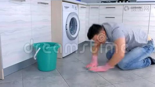 不高兴的疲惫的男人戴着橡胶手套，在厨房里用力地洗和摩擦地板，坐在地板上，侧视。视频