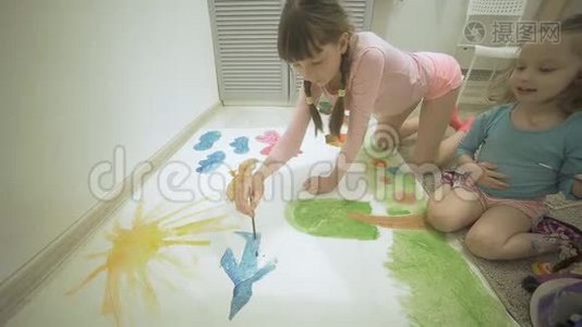 两个女孩在地板上用水彩颜料和画笔在一张大画纸上画画，孩子们`共同的创造力。视频