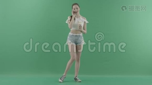 年轻健康的女性在绿色背景下喝鸡尾酒时，会通过智能手机打电话视频