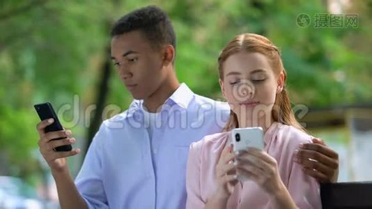 拥抱公园里坐在长椅上的多民族青少年夫妇，聊天的智能手机视频