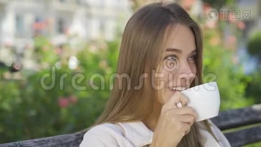 肖像微笑的年轻女子坐在公园的长凳上喝咖啡或茶。 自信的女孩享受美丽视频