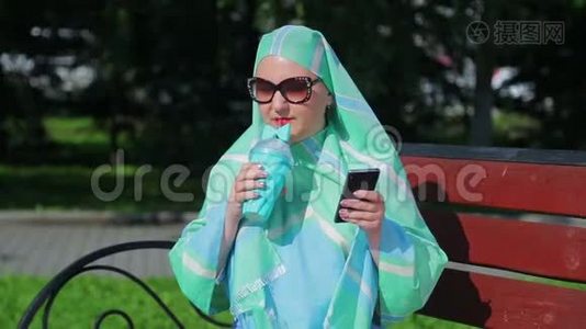 一位戴着浅色围巾和太阳镜的年轻穆斯林妇女坐在公园的长凳上，喝着咖啡，说话视频