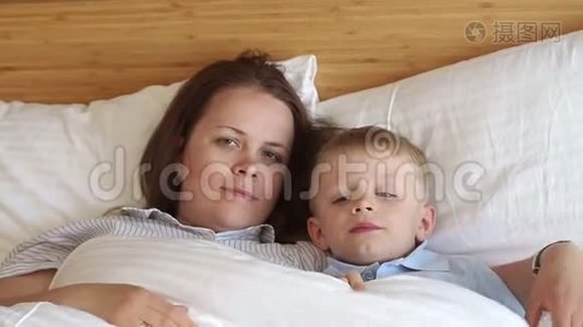 睡意朦胧的妈妈和儿子躺在卧室的床上的白色枕头上。视频