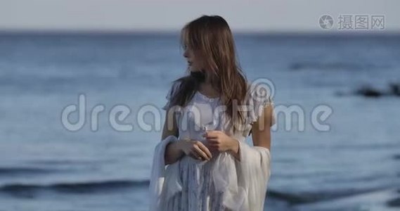 布鲁内特白种人女孩穿着浅色的白色连衣裙站在海浪的背景上，看着一边。 美丽美丽视频