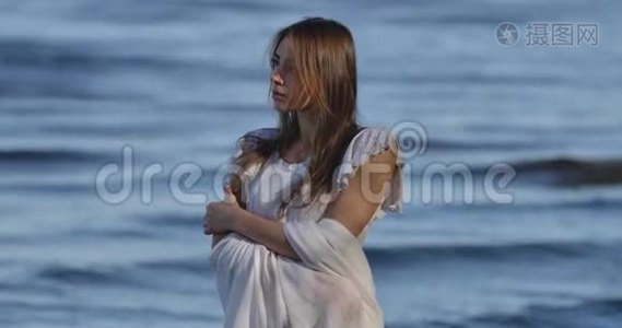 神秘的白种人女孩穿着浅色的白色连衣裙站在海浪的背景上。 美丽的女人看着视频