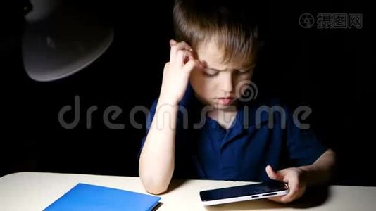 一个孩子坐在家里的桌子旁，晚上用智能手机沉思地看着一幅漫画视频