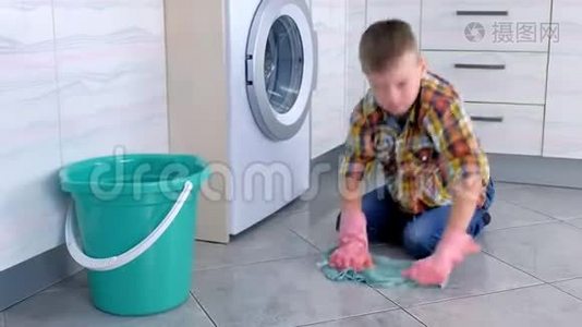穿着橡胶手套的疲惫男孩在厨房里洗地板，看着相机。 儿童`的家庭责任。视频