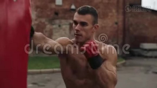 肌肉男拳击手练习拳打脚踢的近侧视图，拳袋在户外。 棕色墙壁视频