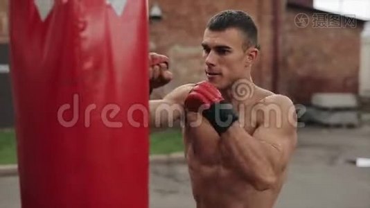 肌肉男拳击手练习拳打脚踢的近侧视图，拳袋在户外。 棕色墙壁视频