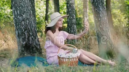 野餐时间。 年轻漂亮的女孩，戴着圆帽子，坐在格子上，摆出自拍的姿势。 公园的户外活动视频