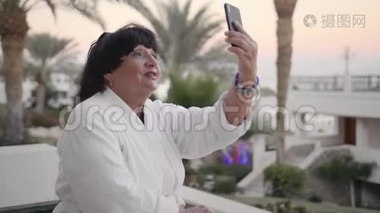 穿着白色特里长袍的快乐白种人高级女性正在一个热带度假胜地休息，并打了一个视频电话。 使用视频