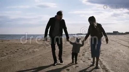 现代，年轻的父母带着儿子走在中间，正走在秋天，春天的海边。 把手举起来视频