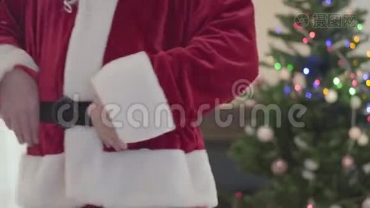 穿着圣诞老人服装的白人老人抱着他的肚子在圣诞树前跳舞。 幸福的概念视频