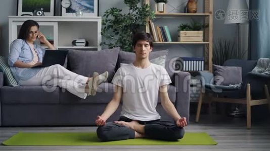 当男朋友在瑜伽垫上冥想时，女孩用手提电脑在手机上说话视频