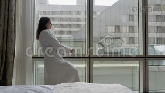 穿着白色浴袍的年轻女子站在酒店公寓的窗户旁，手里拿着智能手机和一杯咖啡视频