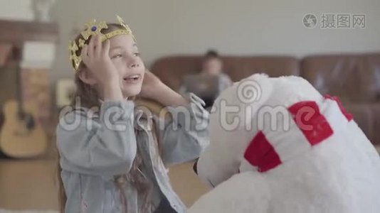 可爱的女孩在玩北方熊玩具，给他戴上皇冠，她的孪生兄弟在沙发上玩手提电脑视频