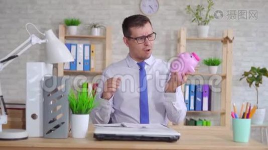 穿着衬衫的男人坐在办公室的笔记本电脑旁，手里拿着一个储蓄罐视频
