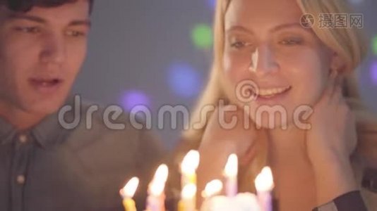特写肖像可爱的微笑金发女孩戴着生日帽吹灭蛋糕上的蜡烛，年轻人坐在附近视频