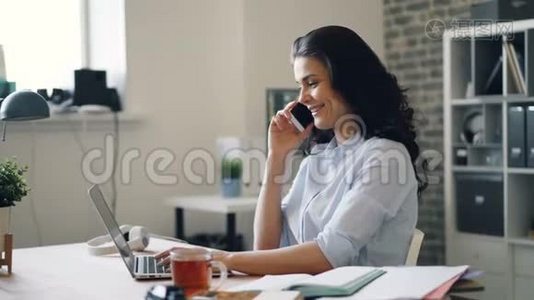 快乐的女员工在手机上说着笑，在办公室里用笔记本电脑视频