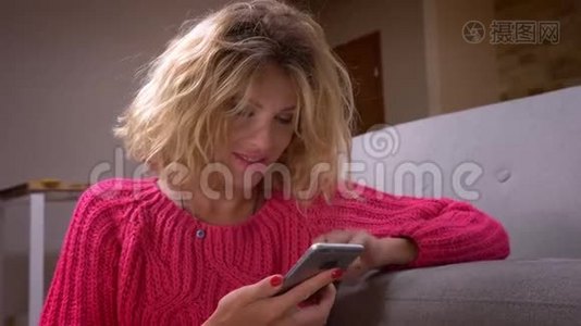 特写镜头，漂亮的家庭主妇穿着粉红色毛衣，在智能手机上冲浪，在家庭氛围中情绪反应。视频
