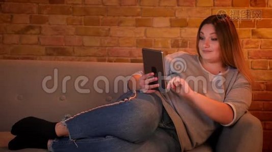 在舒适的家庭氛围中，轻松加上大小的长发模特躺在沙发上愉快地在平板电脑上聊天。视频