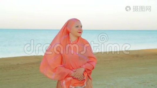 一个穿着橙色披肩的年轻穆斯林女子在沙滩上站在大海的背景下。视频