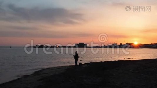 孤独男人的剪影，带着钓鱼竿站在空荡荡的海滩上，地平线上有粉红色的日落。 日落时分钓鱼人剪影视频