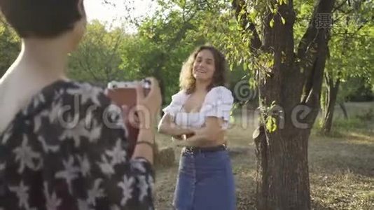 白人女性，留着卷发，在背景中摆姿势，而她的朋友在前景中使用旧相机拍照视频