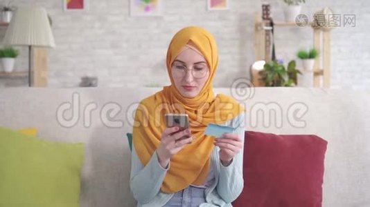 戴着头巾、拿着智能手机、坐在客厅沙发上的银行卡的年轻穆斯林妇女的不幸和烦恼的肖像视频