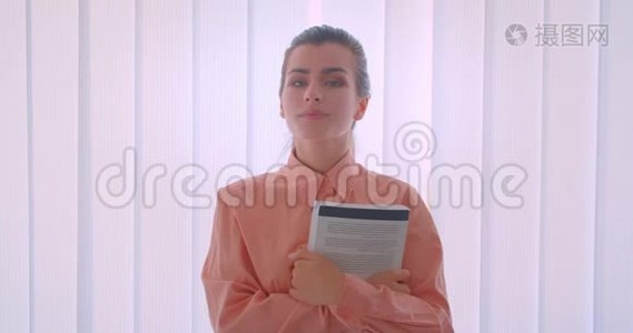 一位年轻有魅力的女商人举着一本书，看着办公室里站在室内的照相机的特写画像视频