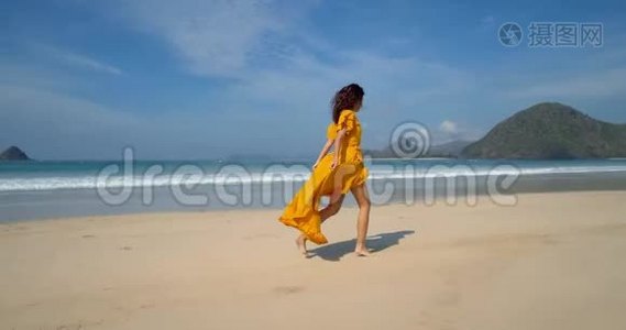 年轻快乐的黑发女郎穿着黄色的裙子，沿着沙滩奔跑，呼吸新鲜空气，欣赏着令人叹为观止的海景。视频