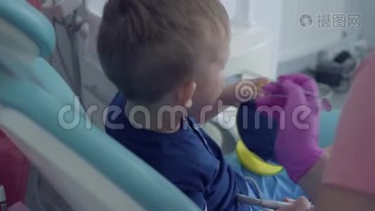 爱的牙医用双手检查坐在椅子上的无忧无虑的小男孩的牙齿，给他看一张镜子特写视频