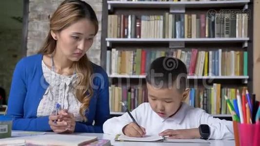 年轻的母亲帮助儿子学习，女人和孩子说话，坐在桌子后面，亚洲孩子做作业，看书视频