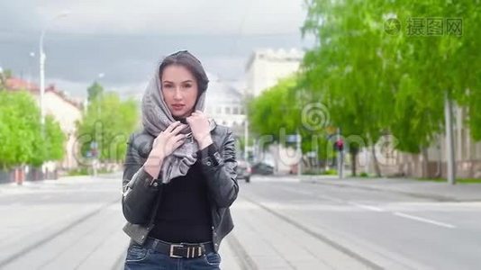 城市背景下穿黑色皮夹克的性感女孩的肖像。 手拿着灰色的围巾戴在头上。 夏日阳光明媚视频