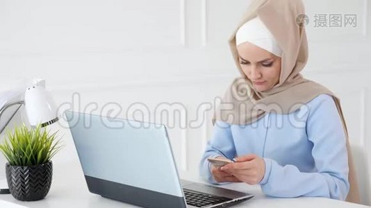 戴头巾的女人正在用手提电脑坐在工作场所看手机上的东西。视频