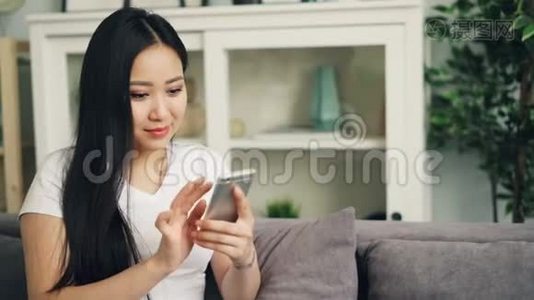 漂亮的亚洲女性穿着白色T恤，正在使用智能手机触摸屏幕，微笑和笑着查看社交媒体视频