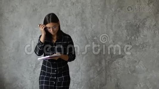 一个年轻快乐的女孩在一个灰色的墙上写笔记本。 时髦的年轻女子视频