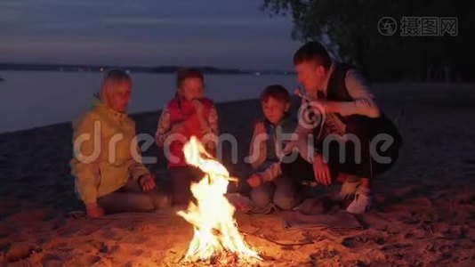 在夜间海滩野餐时，一家人在火上吃饭和聊天视频