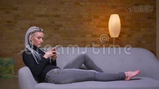 特写侧景拍摄年轻迷人的白种人潮女使用手机躺在室内沙发上视频