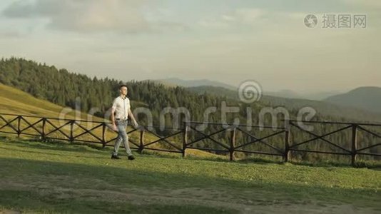 年轻人站在山附近的田野里。 在他身后吃草的马。 日落。 年轻人走在整齐的篱笆上视频