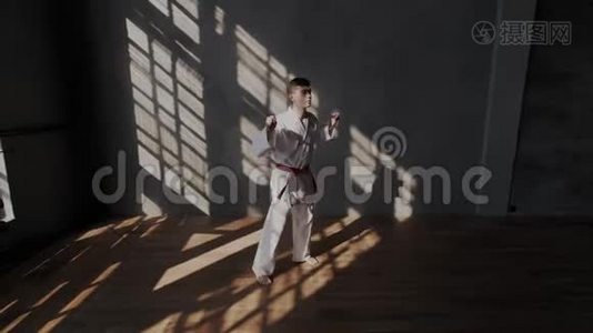 一位年轻的运动员正在准备跆拳道比赛。 带着红腰带的白色和服，光着脚，阳光从窗户进来视频
