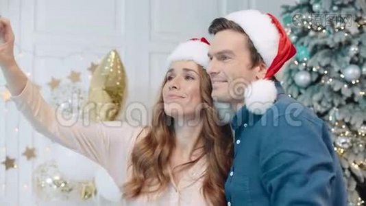 微笑的夫妇在圣诞树附近的圣诞老人帽子里自拍。视频