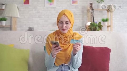 肖像积极的年轻穆斯林妇女戴着头巾，微笑着拿着智能手机和银行卡坐在客厅的沙发上。视频