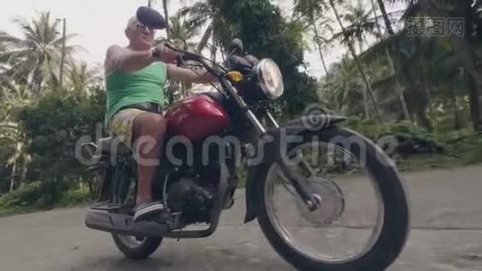 在阳光明媚的日子里，养老金领取者骑摩托车在热带村庄。 骑摩托车在热带行驶的老人摩托车视频