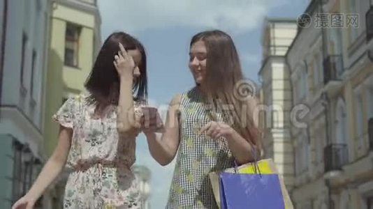 两位快乐的女朋友购物后，拿着购物袋在漂亮的楼房前谈论新闻看着视频