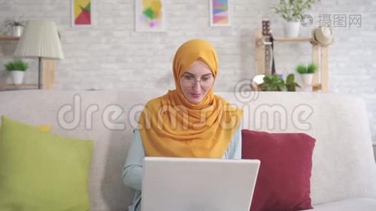 积极的年轻穆斯林妇女，手提电脑坐在客厅的沙发上，看着相机微笑使用银行卡视频