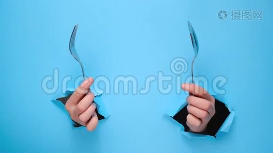 女性手拿着勺子和叉子穿过撕裂的蓝色纸墙。 吃饭的手势。视频