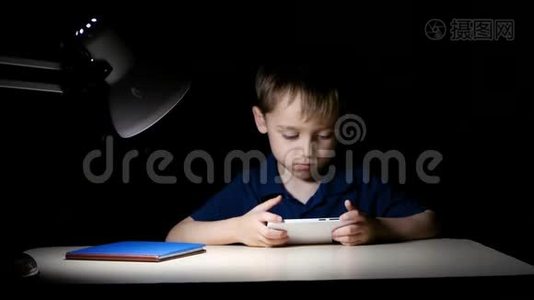 一个孩子晚上在家看卡通片，他用智能手机坐在桌子旁，被一盏灯照亮视频